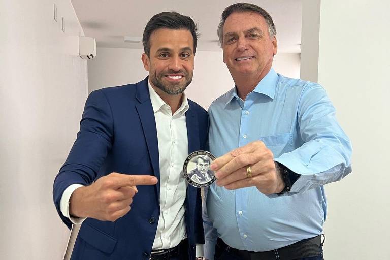 Duas pessoas estão em um ambiente interno, uma delas vestindo um terno azul e a outra uma camisa azul clara. Ambas estão sorrindo e segurando uma medalha, apontando para ela. O fundo é de paredes brancas e há uma iluminação clara