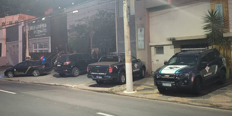 A imagem mostra uma cena noturna com vários veículos de polícia estacionados em frente a um edifício. Há um carro preto com a inscrição 'FEDERAL' e outros veículos de diferentes modelos, incluindo uma caminhonete