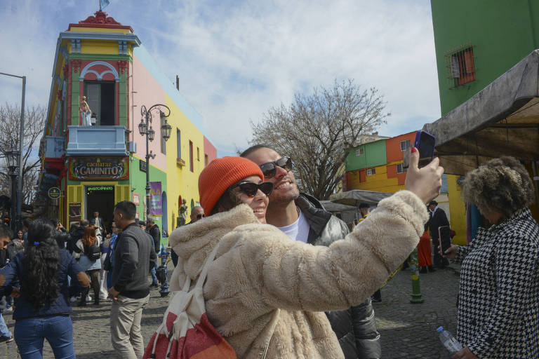 A imagem mostra um casal sorrindo enquanto tira uma selfie em um local vibrante e colorido. Ao fundo, há um edifício com paredes em tons de rosa, amarelo e verde, e uma placa que diz 'cacharrería Caminito'. O casal está vestido com roupas de inverno, a mulher usa um casaco claro e um gorro laranja, enquanto o homem usa uma jaqueta escura. Há várias pessoas ao redor, e o céu está parcialmente nublado.