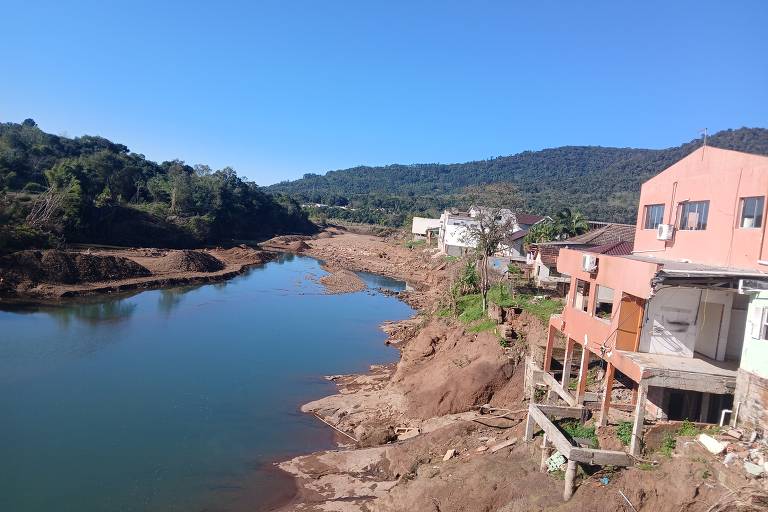 A imagem mostra um ribeirão com águas calmas e margens secas. À direita, há casas em um terreno elevado, algumas com varandas. Ao fundo, há uma vegetação montanhosa e um céu claro e azul. A área ao redor do ribeirão parece ter sofrido erosão, com terra exposta e pouca vegetação.