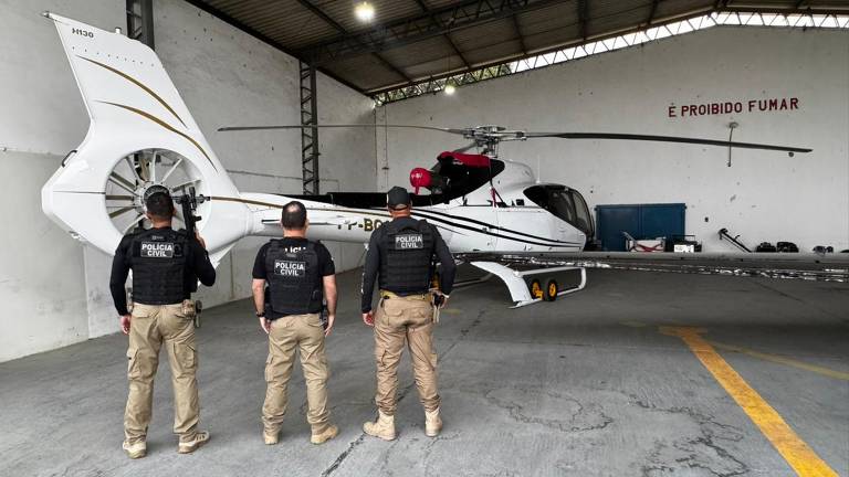 A imagem mostra três policiais civis de costas, em frente a um helicóptero branco com detalhes em dourado. Os policiais estão usando coletes à prova de balas e segurando armas