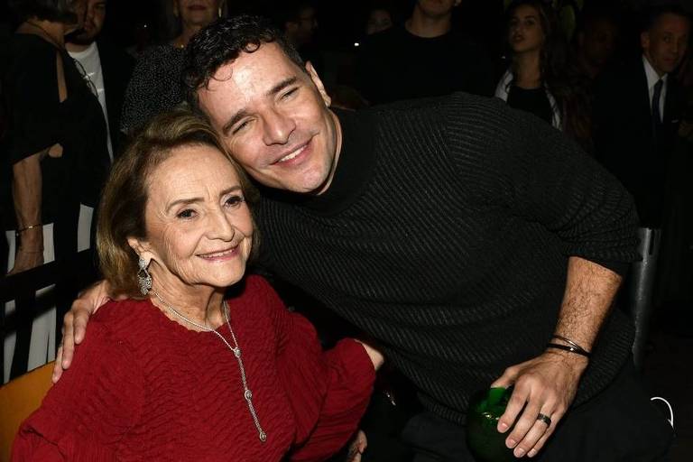 A imagem mostra uma mulher idosa e um homem sorridente em um evento social. A mulher está vestindo um vestido vermelho e segurando um copo com bebida. Ela tem cabelo castanho claro e usa brincos. O homem, que está ao lado dela, usa um suéter preto e também segura um copo com bebida verde. Ambos estão sorrindo e parecem estar se divertindo. Ao fundo, há outras pessoas e uma iluminação suave.