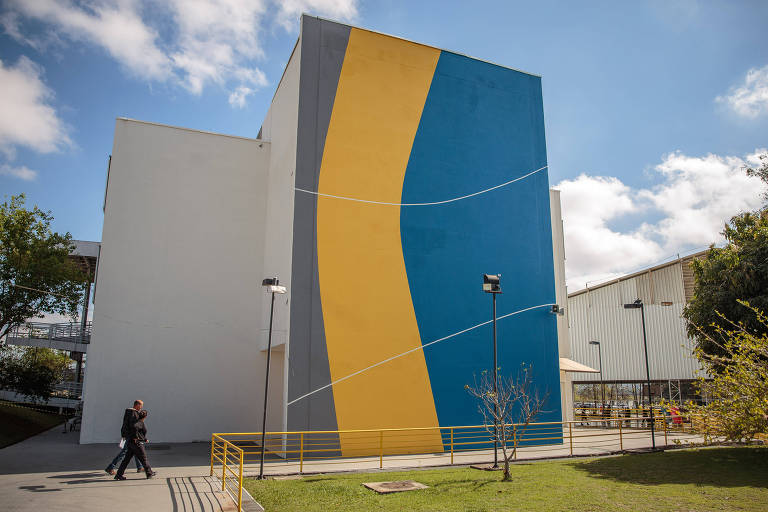 A imagem mostra a fachada de um edifício moderno, pintada em cores azul e amarelo. O edifício tem uma forma retangular e apresenta uma grande parede azul com uma faixa amarela que se curva. No primeiro plano, duas pessoas caminham em direção ao edifício, e há uma área verde com algumas árvores ao redor. O céu está parcialmente nublado.