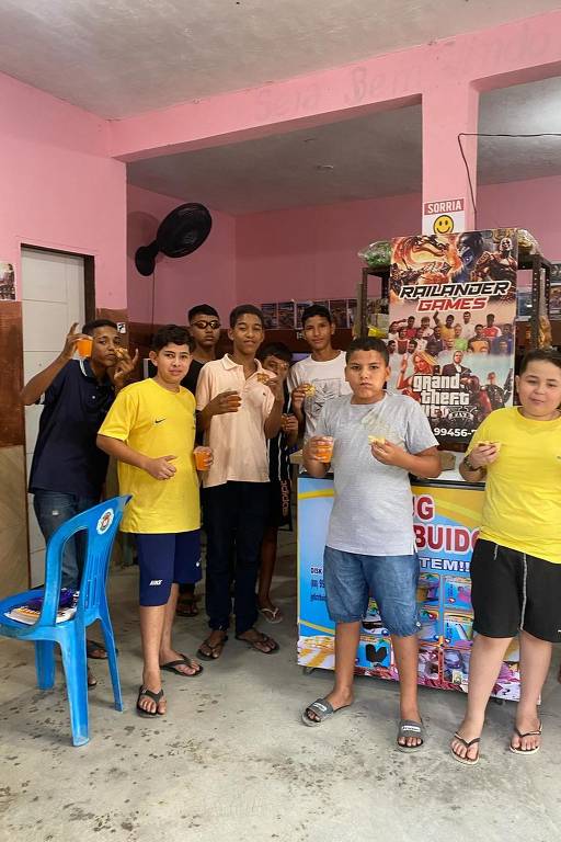 Um grupo de crianças e adolescentes está posando para a foto em um ambiente interno. Eles estão segurando copos com bebidas e alguns têm lanches. O fundo é decorado com pôsteres de jogos e uma mesa com um banner que diz 'RAILANDER GAMES'. As paredes são pintadas de rosa e há uma televisão visível à esquerda.