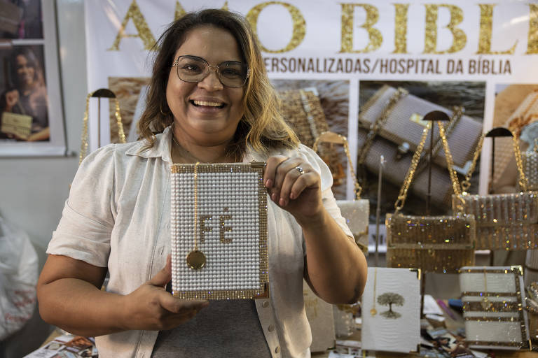 Uma mulher sorridente segura um caderno decorado com pérolas brancas e a palavra 'Fé' em letras douradas. Ao fundo, há uma estande com diversos produtos personalizados, incluindo bolsas e cadernos, todos adornados com detalhes brilhantes. A mulher está vestindo uma camisa clara e óculos, e parece estar em um ambiente de feira ou mercado.