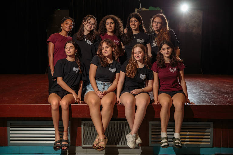 A imagem mostra um grupo de nove jovens mulheres posando em um palco. Elas estão sentadas em uma plataforma, algumas com as pernas cruzadas. As mulheres vestem camisetas pretas e vermelhas, e estão sorrindo. O fundo é escuro, com uma luz brilhante ao fundo, criando um efeito de destaque.