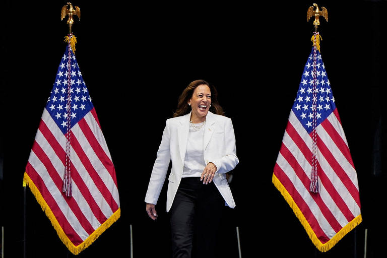 A imagem mostra uma mulher sorridente caminhando em direção à câmera, vestindo um blazer branco e calças pretas. Ela está entre duas bandeiras dos Estados Unidos, que estão posicionadas em ambos os lados. O fundo é escuro, destacando a figura da mulher e as bandeiras.