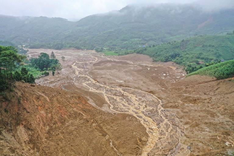 A imagem mostra uma grande área afetada por um deslizamento de terra, com solo exposto e um leito de rio seco visível. Ao fundo, há montanhas cobertas por vegetação e nuvens, indicando um clima nublado. A paisagem é predominantemente marrom, com algumas áreas verdes nas encostas das montanhas.