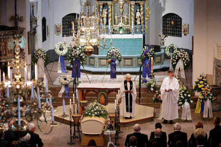 A imagem mostra o interior de uma igreja durante uma cerimônia. No centro, há um altar decorado com flores e um caixão. Dois indivíduos, um vestido de maneira formal e outro com vestes litúrgicas, estão em pé próximos ao altar. Ao fundo, há um grande altar com detalhes ornamentais e várias velas acesas. O ambiente é iluminado e há bancos com pessoas sentadas, observando a cerimônia.