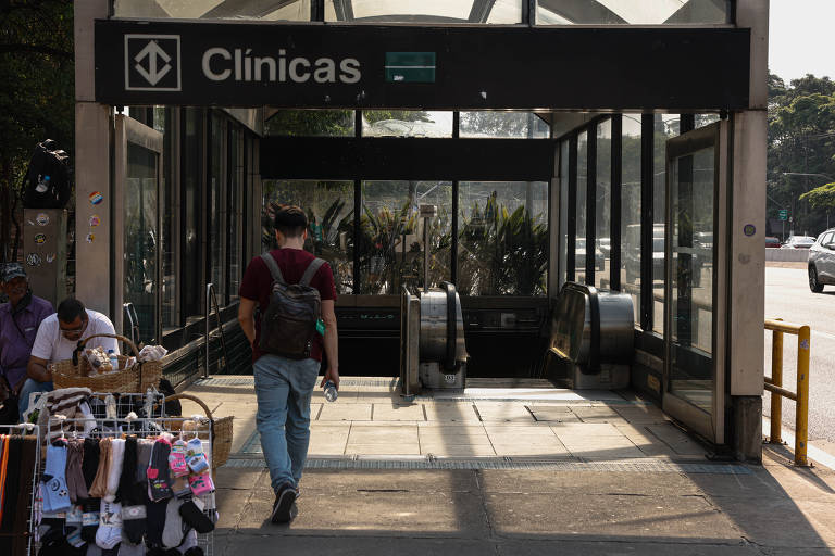 A imagem mostra a entrada da estação de metrô Clínicas. Há um homem caminhando em direção à entrada, que possui escadas rolantes visíveis. À esquerda, há uma banca com produtos expostos, como meias e acessórios. O ambiente é iluminado pelo sol e há árvores ao fundo.