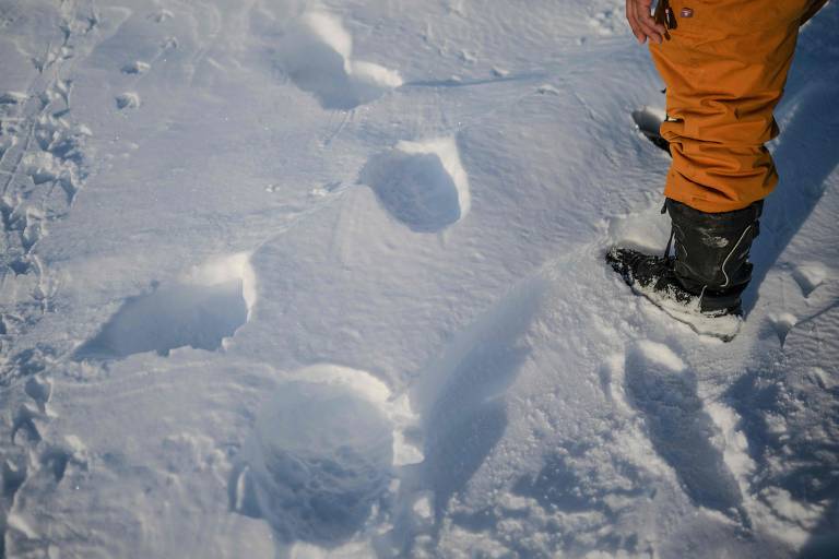 A imagem mostra um pé humano calçado com uma bota preta, posicionado ao lado de várias pegadas na neve. As pegadas são grandes e circulares, com algumas marcas mais profundas, indicando que algo pesado passou por ali. O fundo é coberto por uma camada uniforme de neve branca.