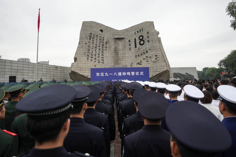 A imagem mostra uma cerimônia com um grande grupo de pessoas em uniformes, incluindo militares e policiais, em frente a um monumento. O monumento é uma estrutura grande e moderna, com inscrições visíveis. Há uma faixa azul com texto em caracteres chineses. Ao fundo, pode-se ver uma bandeira vermelha e árvores.