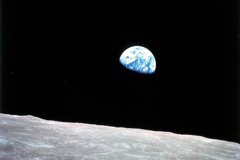 A imagem mostra a Terra vista do espaço, com a superfície lunar em primeiro plano. A Terra aparece como um globo azul e branco, enquanto o fundo é predominantemente negro, representando o espaço. A superfície lunar é acinzentada e apresenta uma textura irregular.