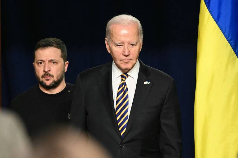 A imagem mostra dois homens em um evento. Um deles está à frente, vestindo um terno escuro e gravata listrada, com uma expressão séria. O outro homem, que está um pouco atrás, usa uma camiseta preta e parece estar atento. Ao fundo, há uma bandeira da Ucrânia com as cores azul e amarelo