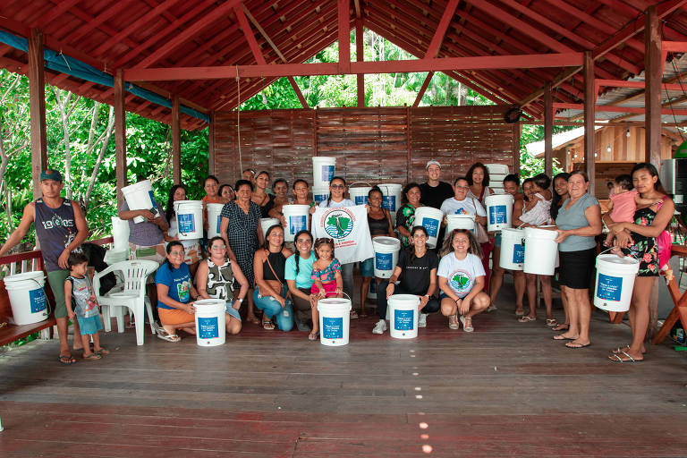 Uma grande reunião de mulheres e crianças em um espaço coberto, segurando baldes brancos com rótulos azuis. O ambiente é cercado por vegetação verde e possui um teto de madeira vermelha. As mulheres estão sorrindo e algumas crianças estão ao lado delas. Há também um homem à esquerda, e todos parecem participar de uma atividade comunitária.