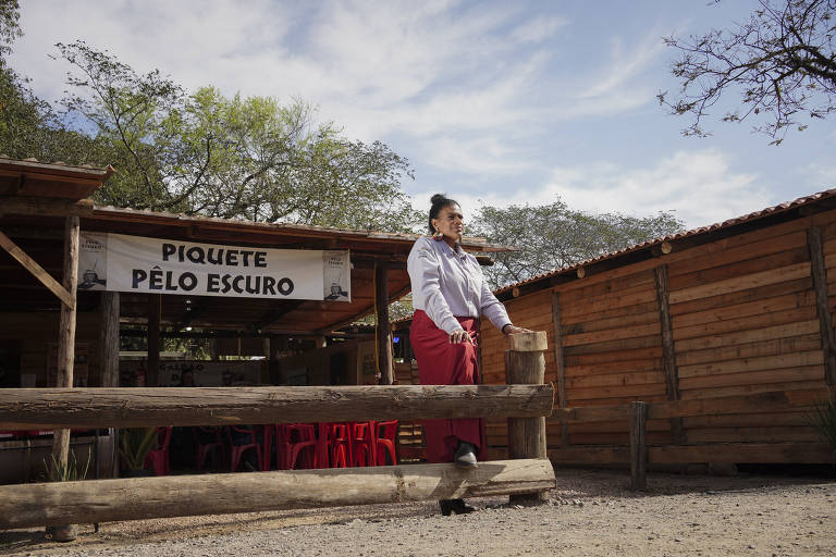 Na foto, uma mulher de blusa clara e saia vermelha está de pé em frente a um letreiro que diz Piquete Pêlo Escuro