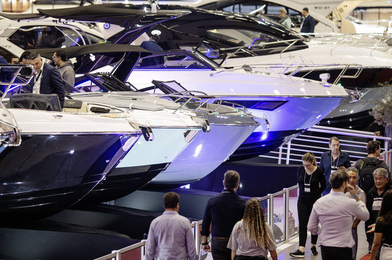 A imagem mostra uma feira de iates, com vários barcos de luxo expostos. Há pessoas caminhando entre os iates, observando e interagindo. Os barcos têm diferentes designs e cores, predominantemente em tons de preto e branco, com iluminação que destaca suas características. O ambiente parece ser um espaço fechado, possivelmente um centro de exposições.