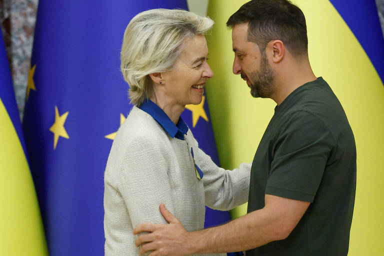 A imagem mostra duas pessoas se cumprimentando em um ambiente formal. Ao fundo, há bandeiras da União Europeia e da Ucrânia. A mulher, vestindo um suéter claro com detalhes em azul, sorri enquanto se inclina em direção ao homem, que está usando uma camiseta escura. Ambos parecem estar em um momento amigável e de cordialidade.