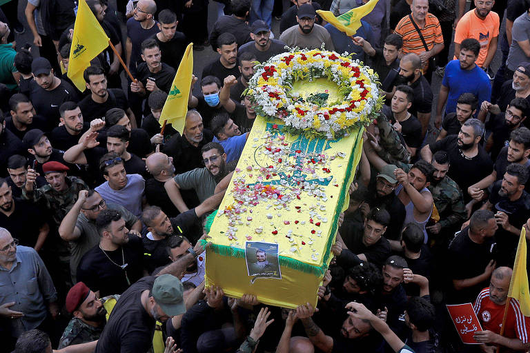 A imagem mostra um grupo de pessoas carregando um caixão decorado com flores e bandeiras. O caixão é predominantemente amarelo e verde, e está cercado por uma multidão que levanta bandeiras. A atmosfera parece ser de luto e homenagem