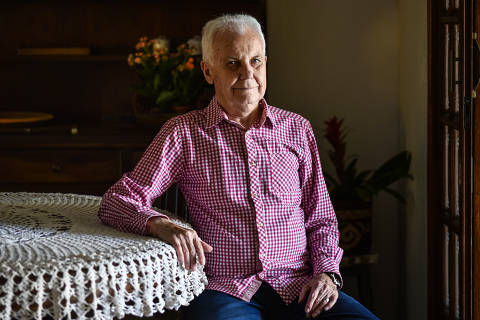 Um homem idoso está sentado em uma cadeira de madeira, com um fundo de plantas verdes e flores rosas. Ele usa uma camisa xadrez em tons de rosa e branco e jeans escuros. O homem tem cabelo grisalho e um sorriso suave.