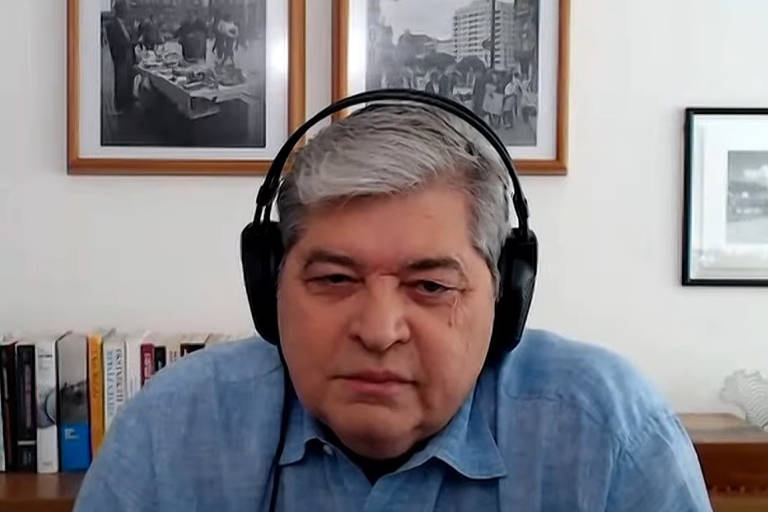 Print mostra o busto de Datena, um homem branco de cabelos grisalhos, usando uma camisa azul clara e fones de ouvido pretos. Ao fundo, há duas fotos emolduradas na parede e uma estante com livros.