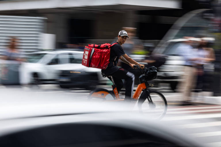 A imagem mostra um entregador em uma bicicleta, com uma mochila vermelha de entrega nas costas, passando rapidamente por uma rua movimentada. Ao fundo, há carros e pedestres, com um leve desfoque que sugere movimento.