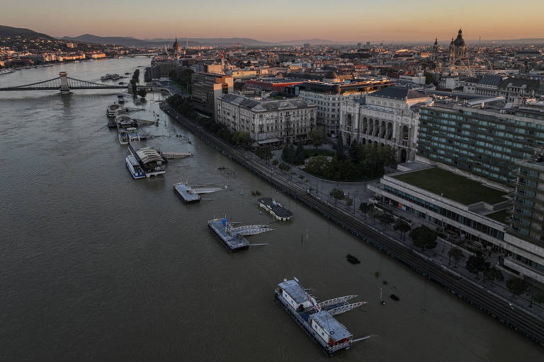 A imagem mostra uma vista aérea do Rio Danúbio em Budapeste, com vários barcos ancorados na água. À margem do rio, há edifícios e áreas urbanas, incluindo um grande edifício à direita. O céu apresenta um tom alaranjado, indicando o pôr do sol. A ponte visível conecta as duas margens do rio.