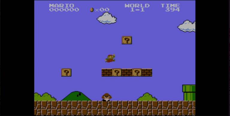 A imagem mostra uma cena do jogo Super Mario Bros. O fundo é azul com algumas nuvens brancas. Há blocos de perguntas flutuando acima de duas colinas verdes. Um tubo verde está à direita, e o personagem principal, Mario, está na parte inferior da tela, próximo a um bloco de perguntas.