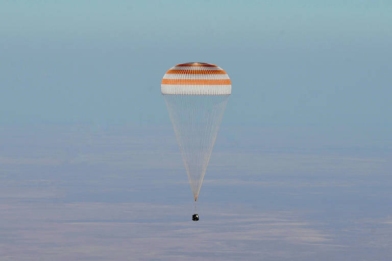 A imagem mostra um paraquedas laranja e branco descendo lentamente em um céu azul claro. Abaixo do paraquedas, há um objeto escuro que parece ser uma cápsula espacial, enquanto o fundo é composto por uma superfície plana e clara.