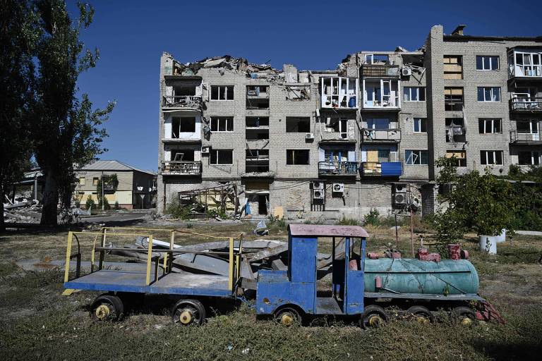 A imagem mostra um edifício em ruínas, com várias janelas quebradas e partes da estrutura desmoronadas. Em primeiro plano, há um brinquedo de trem de cor azul e amarelo, estacionado em um terreno vazio, ao lado de um tanque de água. O céu está limpo e azul, e há algumas árvores ao fundo.