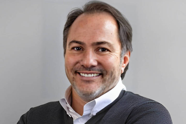 Um homem com cabelo escuro e barba bem aparada, sorrindo. Ele está usando uma camisa branca com um suéter escuro por cima. O fundo é de uma cor neutra, clara.
