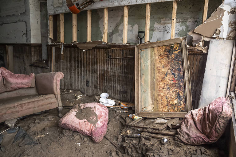 A imagem mostra um ambiente interno em estado de abandono. Há um sofá de cor clara à esquerda, coberto de poeira e sujeira. No chão, há almofadas sujas e um quadro inclinado contra a parede, que parece estar danificado. A parede ao fundo está parcialmente exposta, com tábuas de madeira visíveis. O ambiente parece ter sido afetado por umidade e deterioração