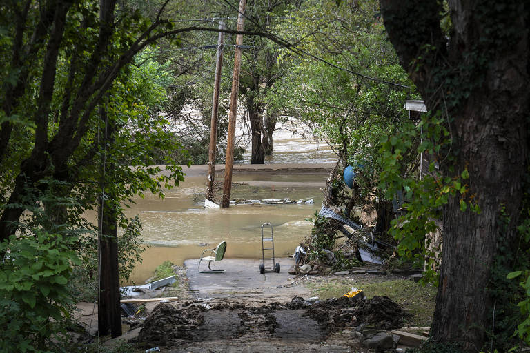 A imagem mostra uma área residencial afetada por uma inundação. O nível da água está alto, cobrindo partes do chão e subindo em direção a postes de eletricidade. Árvores cercam a área, e há detritos visíveis ao longo da margem da água. O céu parece nublado, indicando condições climáticas adversas