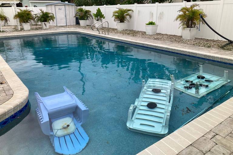 A imagem mostra uma piscina com água clara e tranquila. Dentro da piscina, há três flutuadores coloridos: um azul claro e dois em tons de verde. Ao redor da piscina, há um cercado branco e plantas em vasos, criando um ambiente relaxante.