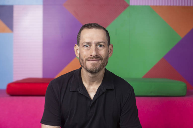 Um homem com cabelo curto e barba está sentado em um ambiente colorido. Ele usa uma camisa preta e sorri para a câmera. Ao fundo, há uma parede com formas geométricas em várias cores, incluindo verde, roxo, laranja e rosa, além de almofadas coloridas em tons de vermelho e verde.