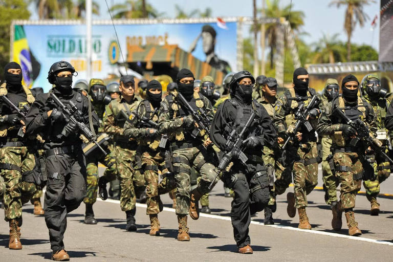 Um grupo de soldados marchando em uma rua, todos vestidos com uniformes camuflados e equipados com armas. Alguns soldados estão usando máscaras e capacetes, enquanto outros estão em uniformes pretos. Ao fundo, há um banner com a imagem de um soldado e a frase 'SOLDADO DO BRASIL'. O ambiente é ensolarado, com palmeiras visíveis ao fundo.