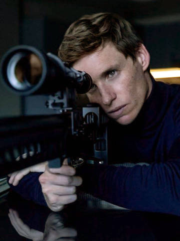 Cena da série O Dia do Chacal  (The Day of the Jackal) com Eddie Redmayne *** Local Caption *** Um homem está posicionado em uma mesa, segurando um rifle com uma mira. Ele tem cabelo curto e castanho claro, e está usando uma camisa de gola alta preta. O ambiente é escuro, com luz suave ao fundo, e há um caderno aberto e uma cápsula de munição na mesa.