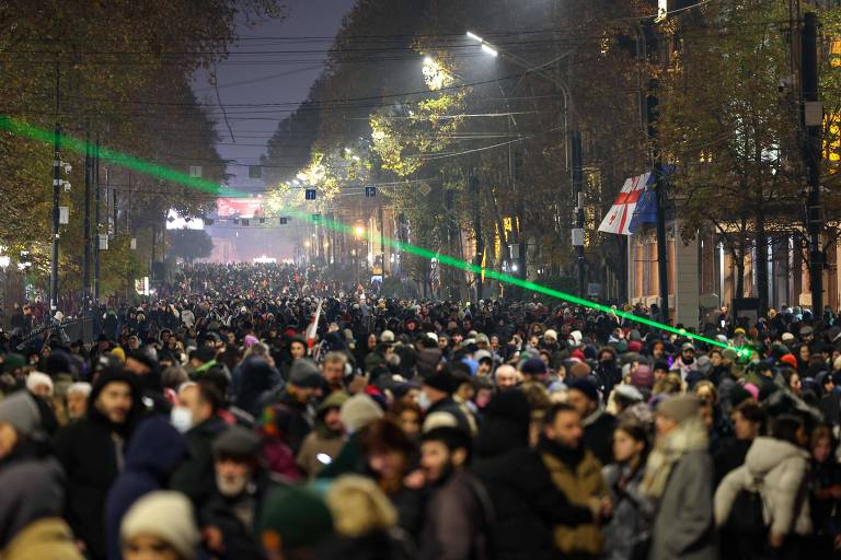 A imagem mostra uma grande multidão reunida em uma rua iluminada à noite. Há árvores ao longo da calçada, e algumas bandeiras estão visíveis ao fundo. Feixes de laser verde cortam o ar. A atmosfera é de protesto, com muitas pessoas vestindo roupas de inverno.