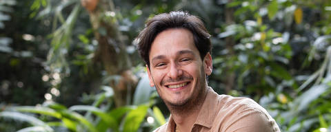 Bruno Luperi (Autor)

 *** Local Caption *** Um homem sorridente está sentado em uma pedra em um ambiente natural, cercado por plantas verdes. Ele usa uma camisa bege e jeans escuros, com tênis claros. O fundo é composto por vegetação densa, sugerindo um cenário de floresta.