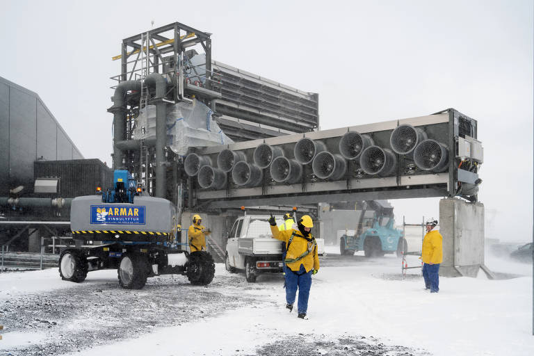 A imagem mostra um ambiente de trabalho em condições de neve intensa. Há uma estrutura grande com vários ventiladores na parte superior, e um veículo de transporte com a marca 'ARMAR' está em primeiro plano. Dois trabalhadores, vestidos com roupas amarelas e azuis, estão na cena, um deles acenando. Ao fundo, há mais veículos e uma construção coberta de neve.