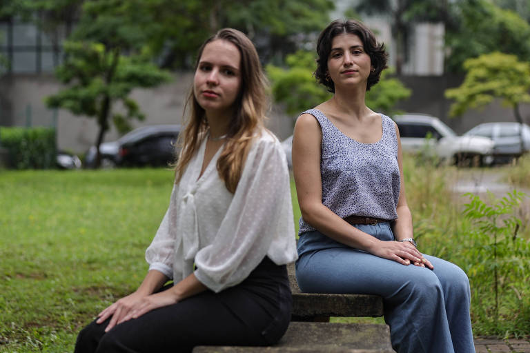 A imagem mostra duas mulheres sentadas em um banco em um parque. A mulher à esquerda tem cabelo longo e liso, vestindo uma blusa branca e calças escuras. A mulher à direita tem cabelo curto e ondulado, usando uma blusa cinza e calças jeans claras. Ao fundo, há árvores e carros estacionados