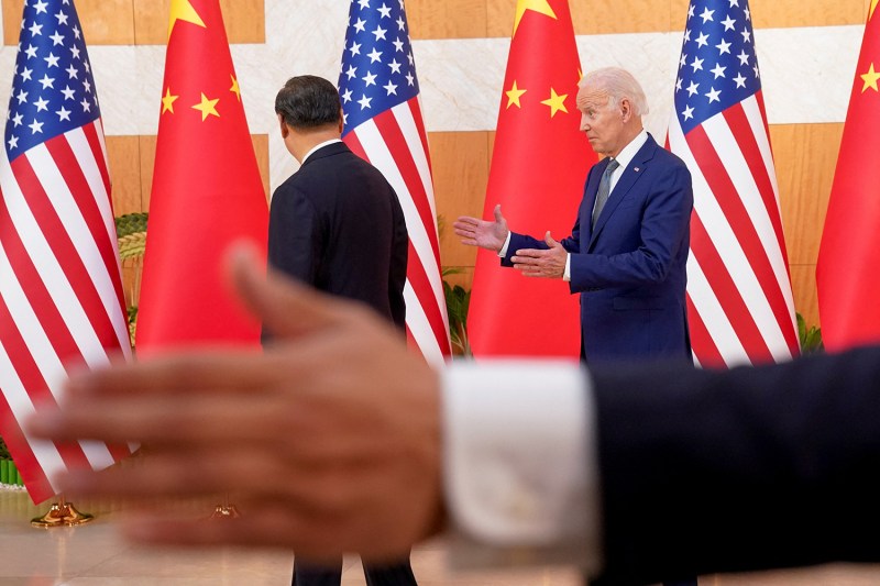 U.S. President Joe Biden, right, gestures toward Chinese President Xi Jinping seen from behind at left. Behind them are U.S. and China flags. A blurry hand gestures to the left in front of them.