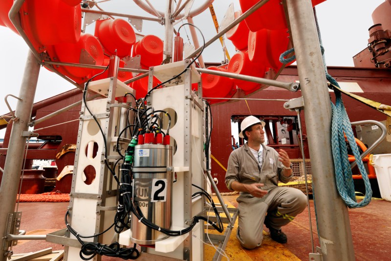 Andrew Sweetman, a deep-sea ecology professor wearing a gray boiler suit and white hard hat, kneels on one knee as he gestures to research equipment on the deck of a ship beneath a pale cloudy sky in the Pacific Ocean.