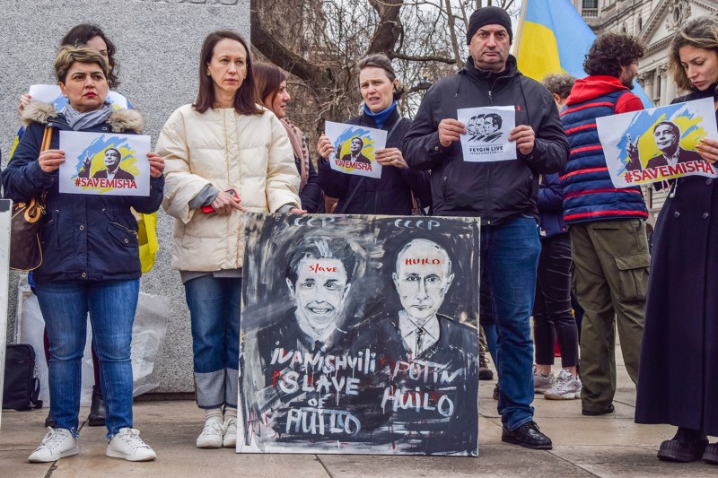 A small group of protesters hold signs in support of Mikheil Saakashvili. In front of them is a larger sign depicting Georgian Prime Minister Bidzina Ivanishvili and Russian President Vladimir Putin.