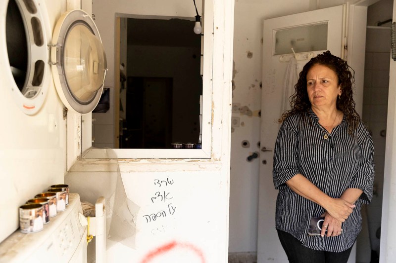 A woman wearing a short sleeve shirt stands in a doorway inside a house. Bullet holes can be seen in the walls behind her and writing and spray paint is on a wall.