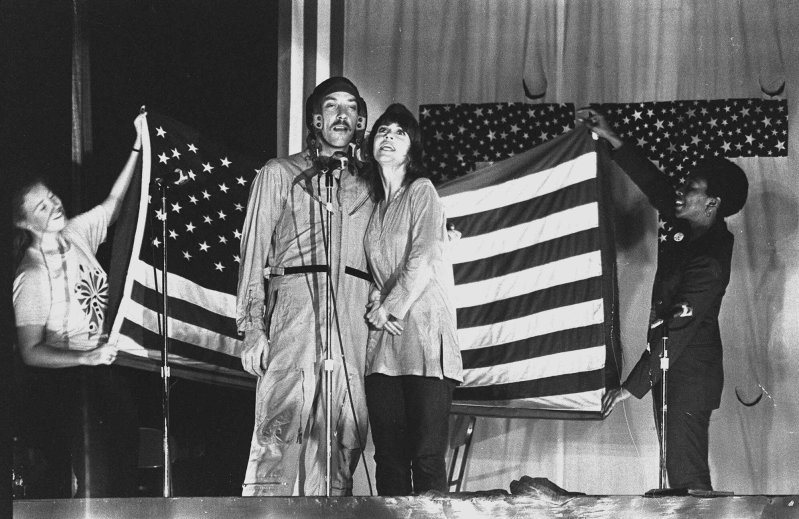 Donald Sutherland and Jane Fonda speak or sing into a microphone on a stage in front of the U.S. flag, held by two smiling women.