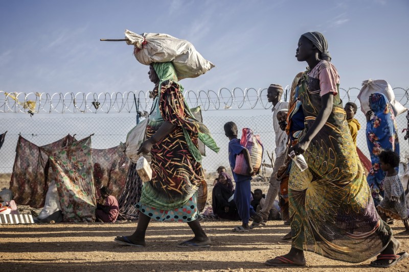 Sudanese refugees walk along a dirt path next to a metal fence topped with barbed wire beneath hazy clouds against a blue sky. Many carry belongings with them, including a woman at the front of the pack balancing a large sack on her head as she carries others in her arms. Leaning against the fence are tents and cloths draped to create shade.