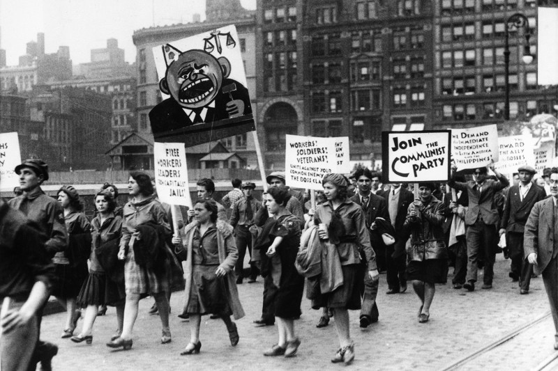 Marchers, most of them women in heels, dresses, coats, and hats carry signs for the communist part as they walk down a street in New York.