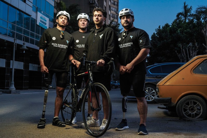 Four men, three with prosthetic legs, wear bike helmets and t-shirts as they stand in a parking lot, gathered around a bicycle held up by the man second from the right. The sky above is a dim, dark blue; it seems to be either dawn or dusk.