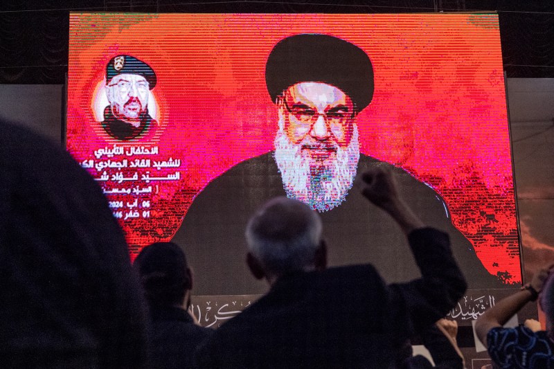 A crowd of people are seen from behind, dimly lit, as they watch a speech from a Hezbollah leader delivered via a large screen. The nearest spectator holds a fist in the air toward the bearded man onscreen, who speaks in front of a bright red background.